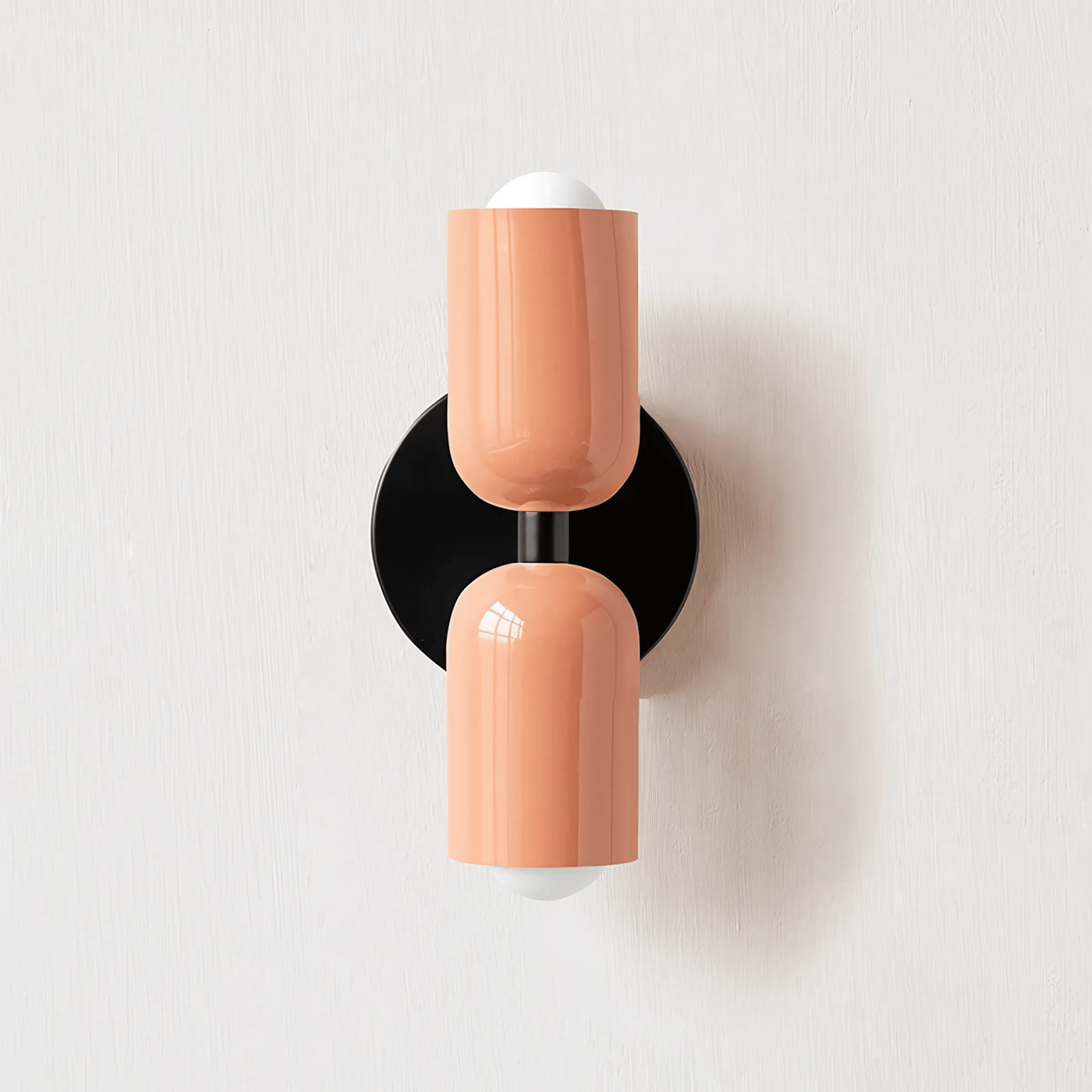 A peach-colored metal wall sconce with a black circular base, mounted on a white textured wall.