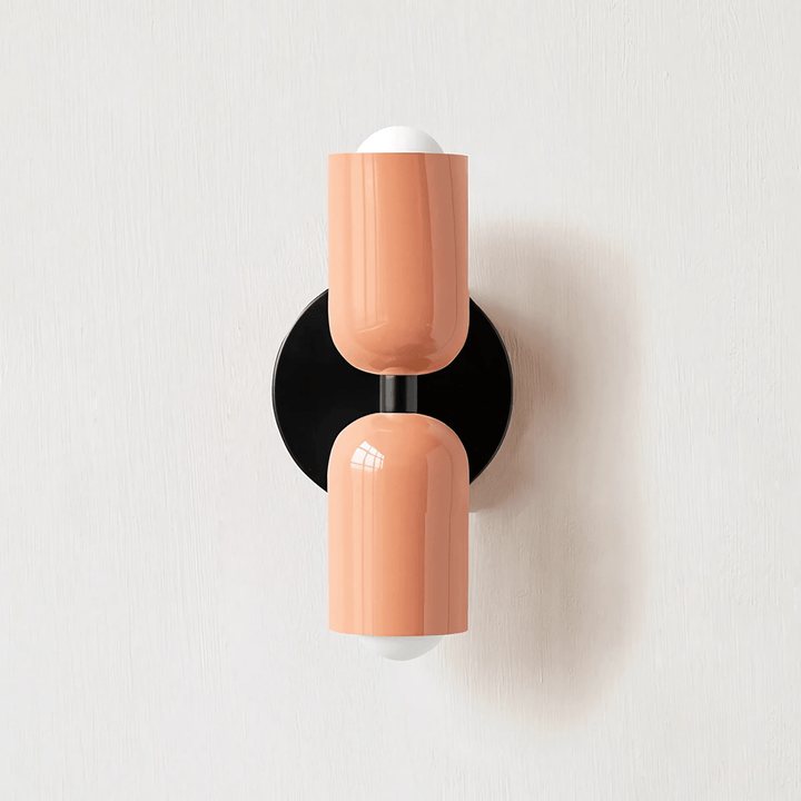 A peach-colored metal wall sconce with a black circular base, mounted on a white textured wall.