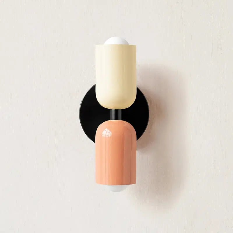 A wall sconce with a black circular base, a cream-colored cylinder, and a peach-colored cylinder.