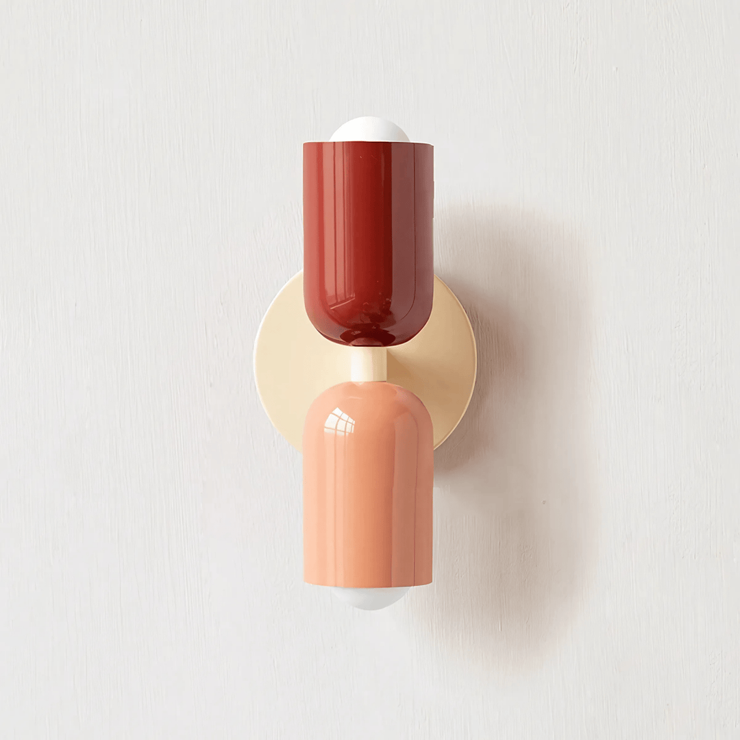 A colorful metal wall sconce with a red top shade and a peach bottom shade, mounted on a cream circular backplate.