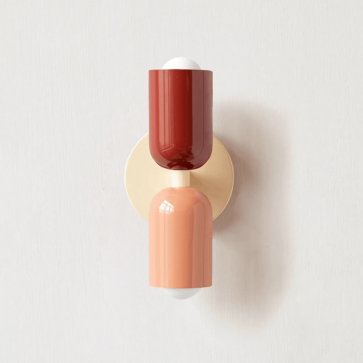 A colorful metal wall sconce with a red top shade and a peach bottom shade, mounted on a cream circular backplate.