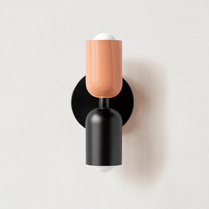 Black and peach metal wall sconce with two cylindrical shades and exposed bulbs.