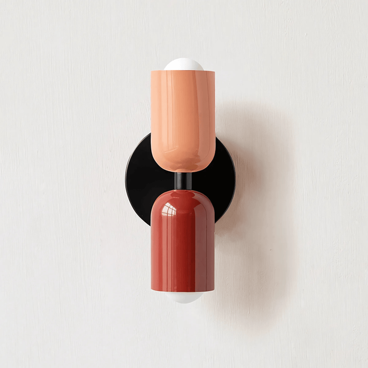 A double-headed metal wall sconce with a black circular base, featuring a coral-colored cylinder above a rust-colored cylinder, each topped with a white bulb.