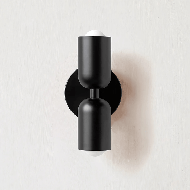 Black metal wall sconce with two cylindrical shades and exposed white bulbs.