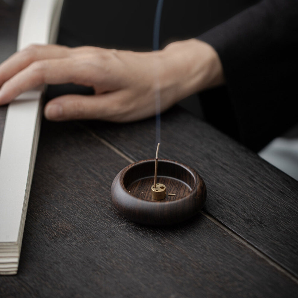 Round solid wood incense holder with a brass insert and a lit incense stick emitting smoke.