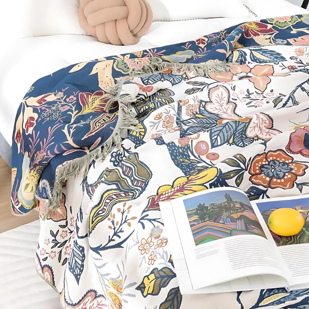 Reversible floral cotton throw blanket with tassels draped on a bed with an open book and an orange.
