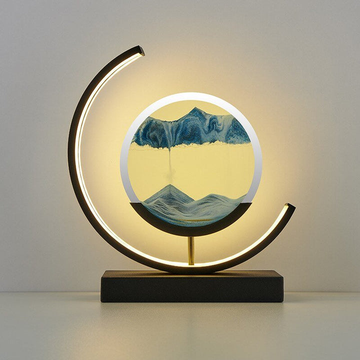 Modern crescent lamp with a circular sand art display showing blue mountains and falling sand.