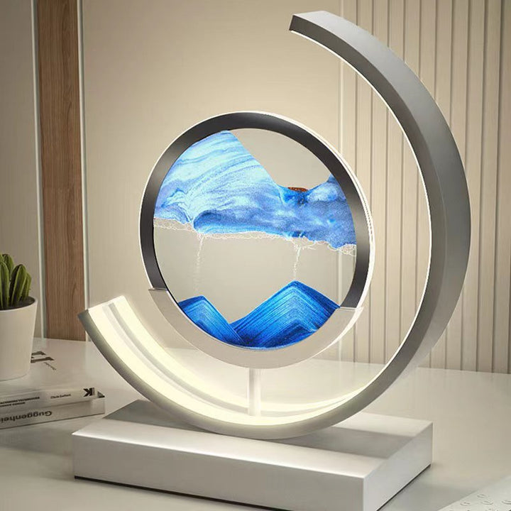 Modern LED table lamp with a circular frame containing flowing blue sand art, illuminated by a curved light.