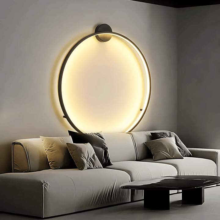 Modern circular wall sconce with warm ambient lighting above a light gray sectional sofa.