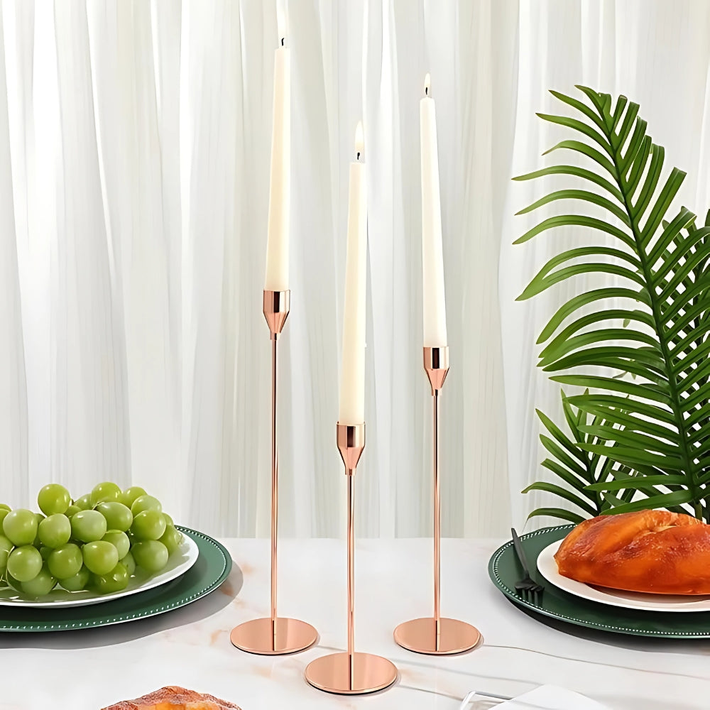 Three tall, slender rose gold stainless steel candle holders with cream candles, set on a table with grapes and bread.