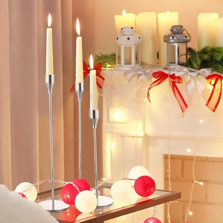 Three tall, slender stainless steel candle holders with cream-colored candles, arranged on a glass table.