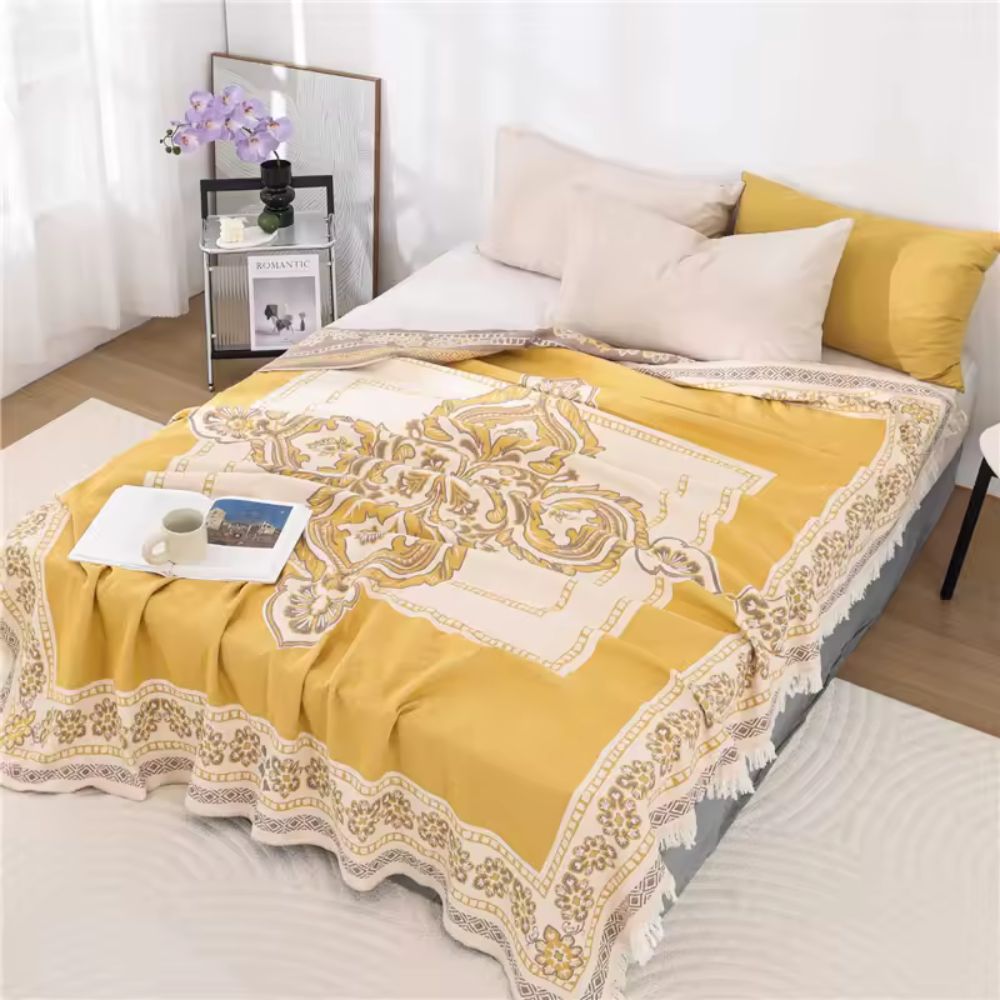 Yellow cotton boho mandala blanket with fringe draped on a bed with pillows and a book.