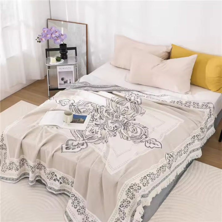 A beige cotton boho mandala blanket with fringe is draped over a bed with pillows.