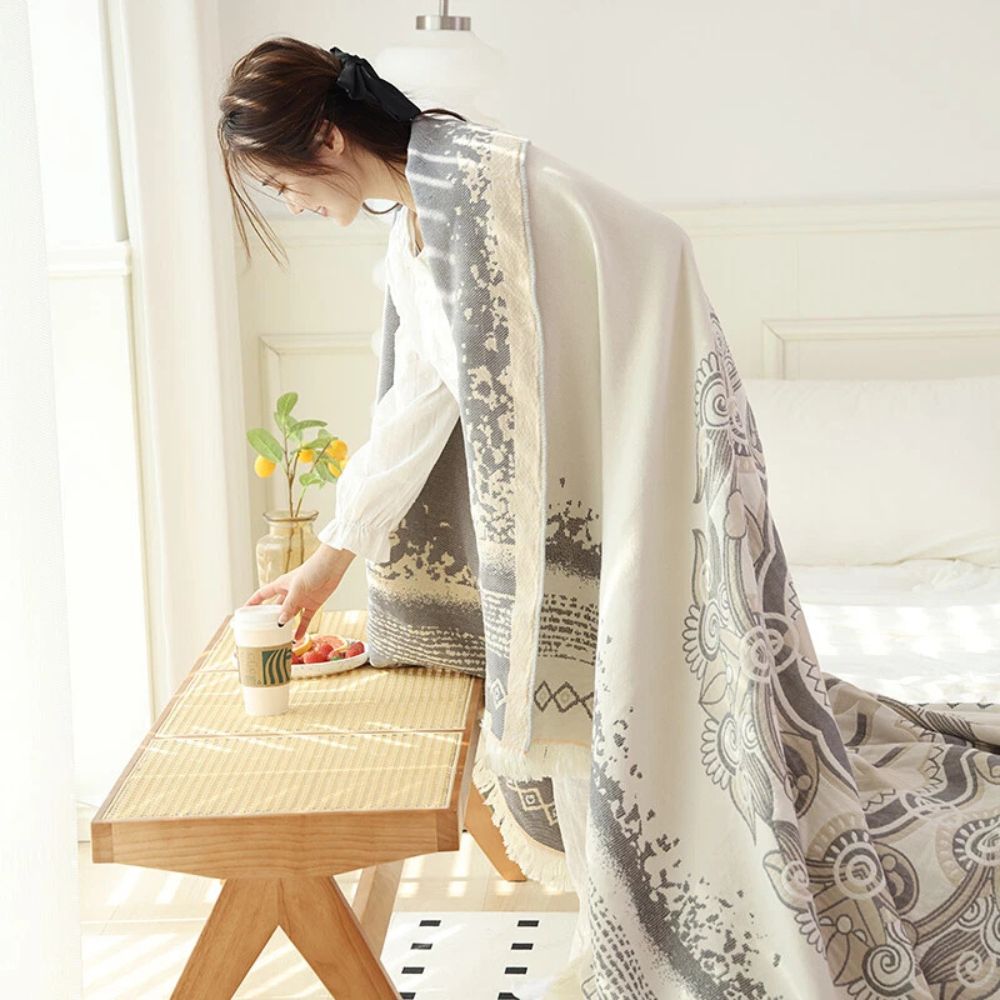 A woman in a white shirt is draped in a white and gray boho mandala patterned cotton blanket, reaching for a coffee cup on a wooden bench.