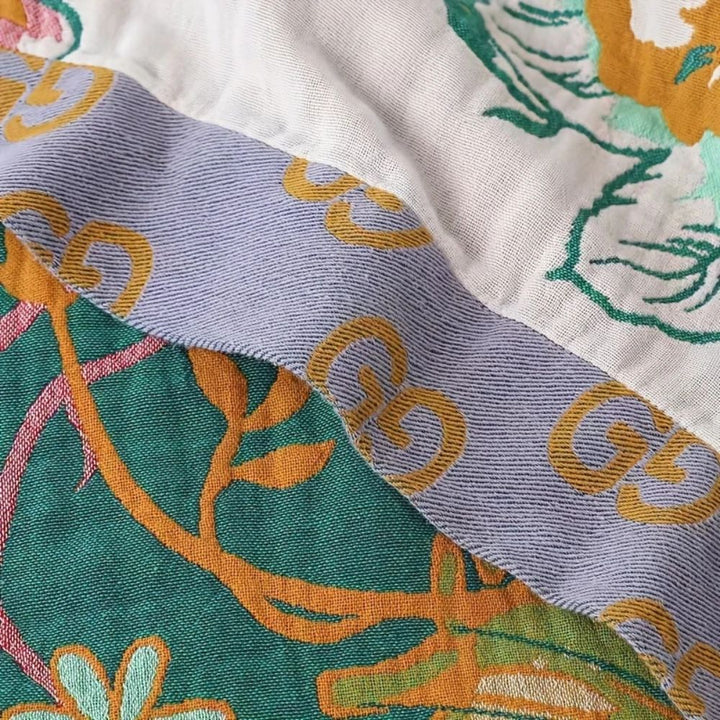 Close-up of a cotton blanket with a pattern of green leaves, orange flowers, and a light purple band with gold interlocking Gs.