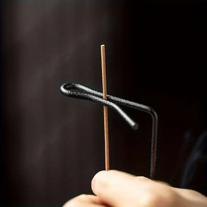 A hand holds a wooden incense stick with a metal holder, with smoke rising against a dark background.