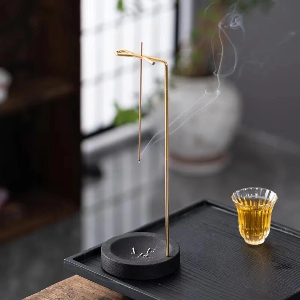 Minimalist wooden incense burner with a thin metal holder and a lit incense stick, smoke rising, next to a glass of tea.
