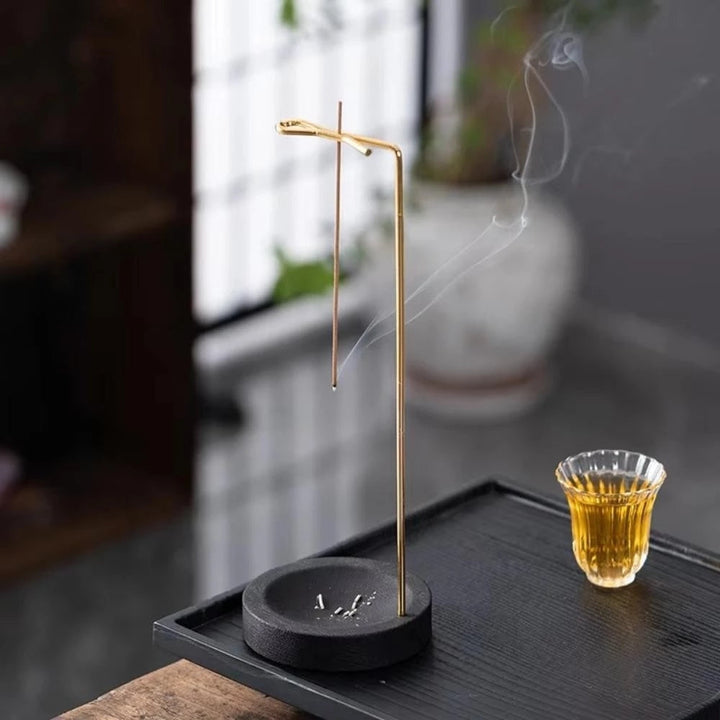 Minimalist wooden incense burner with a thin metal holder and a lit incense stick, smoke rising, next to a glass of tea.