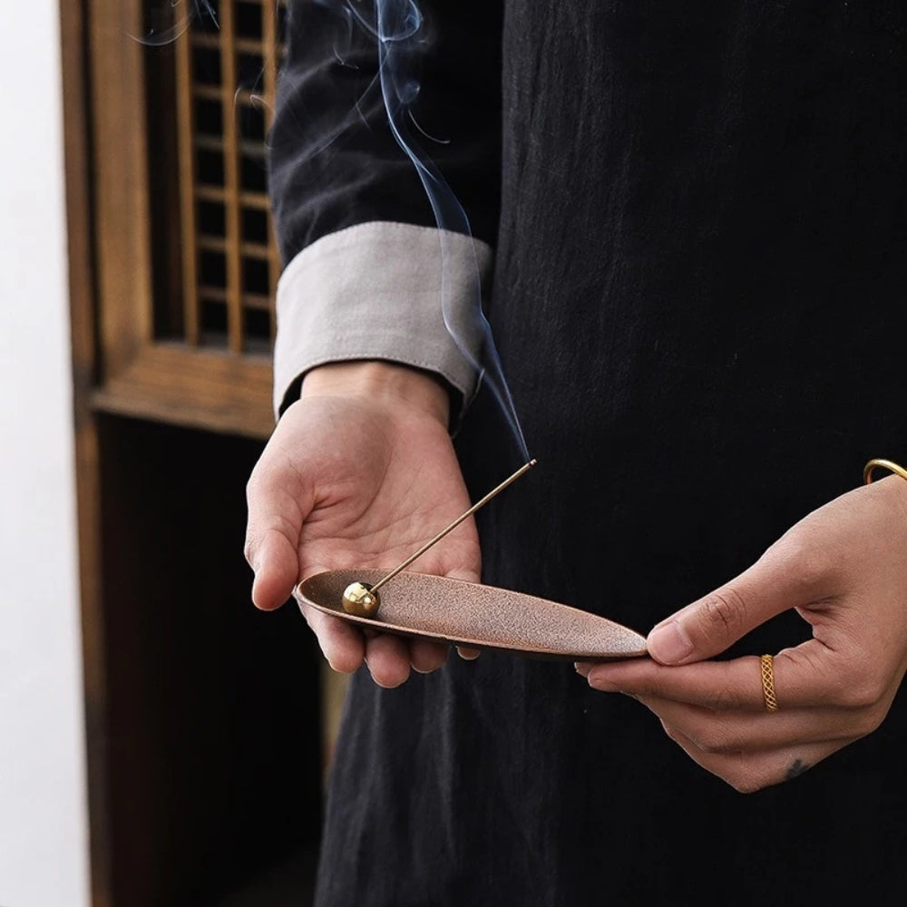 Hands holding a bronze-colored, oval incense holder with a brass bead and a lit incense stick emitting smoke.