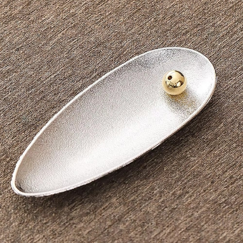 Silver alloy incense holder with a small gold bead on the right side, resting on a textured brown surface.