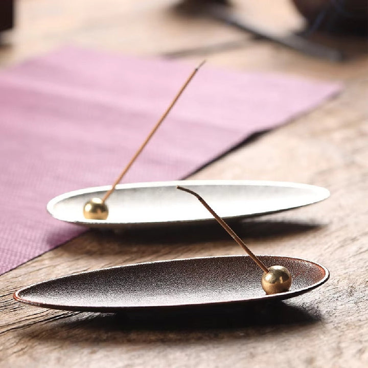 Two oval-shaped incense holders, one silver and one dark brown, each with a brass bead holding an incense stick.