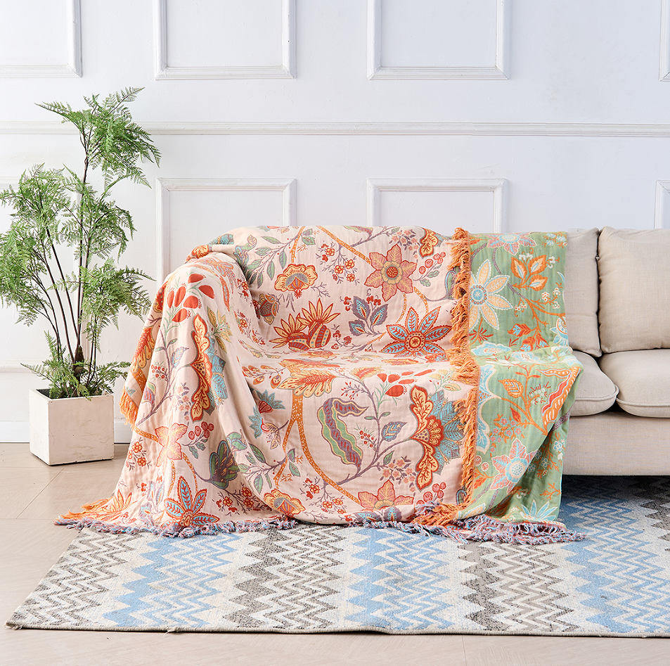 A rustic floral boho cotton blanket draped over a sofa, featuring a beige base with orange, blue, and pink floral patterns and tasseled edges.