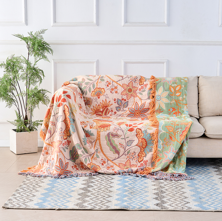 A rustic floral boho cotton blanket draped over a sofa, featuring a beige base with orange, blue, and pink floral patterns and tasseled edges.