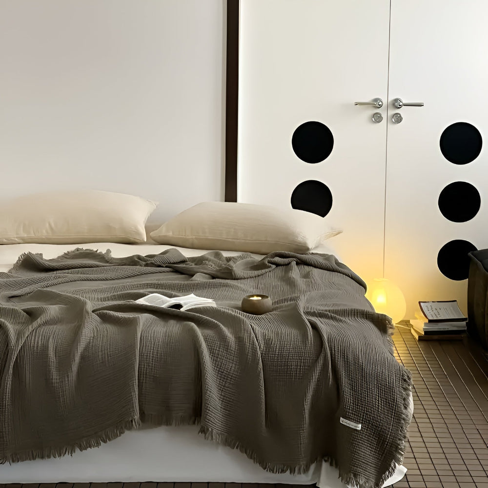 Ash Gray cotton crinkle tassel blanket draped over a bed with two pillows and a book.