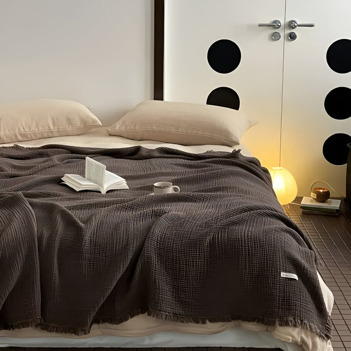 A dark brown cotton crinkle tassel blanket is draped over a bed with two pillows, a book, and a mug.