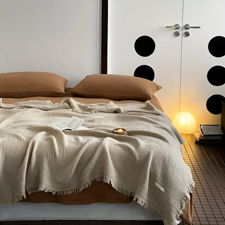 Cream cotton crinkle tassel blanket draped on a bed with brown pillows and a book.