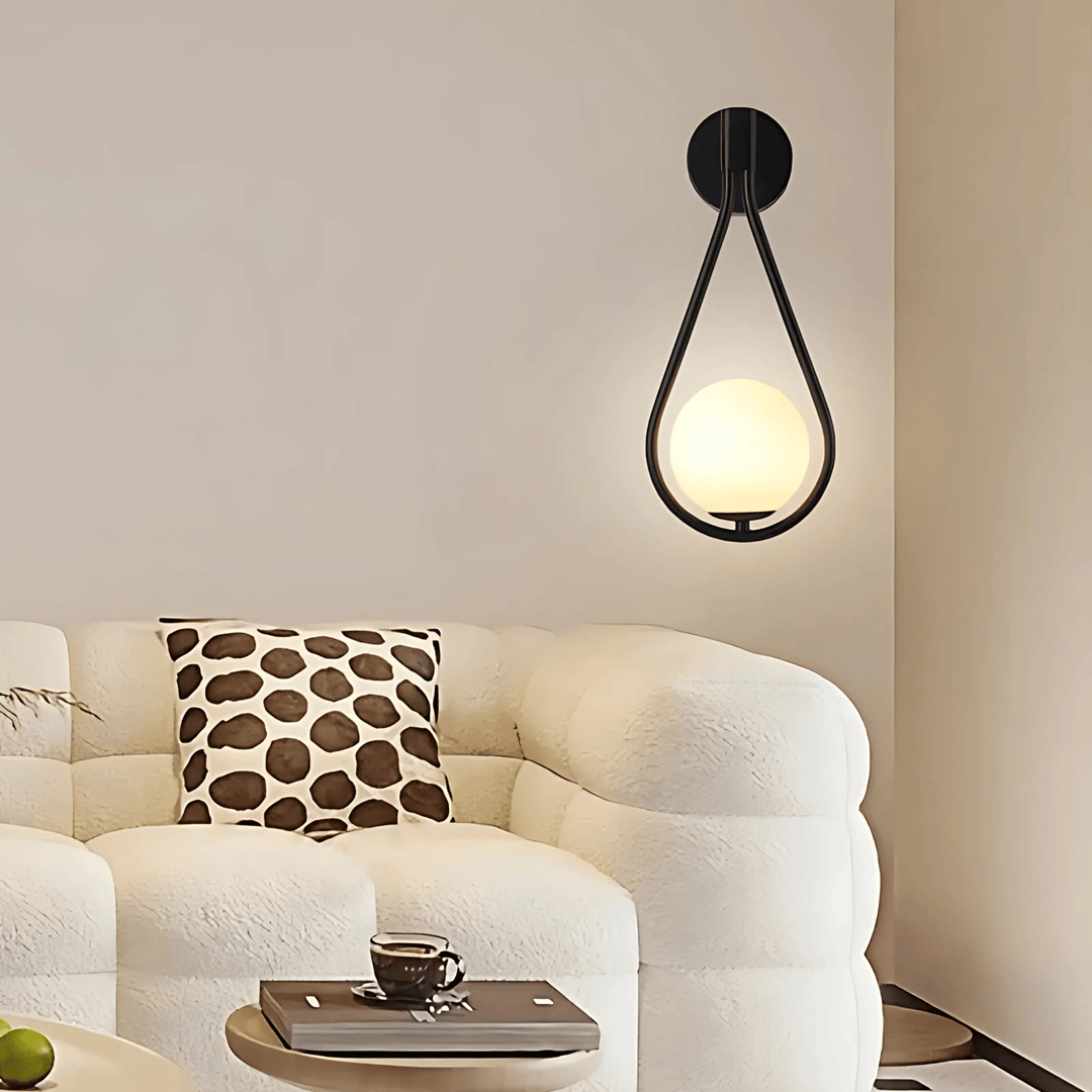 Teardrop wall sconce with a black frame and a glowing white orb, mounted on a light-colored wall above a white sofa.