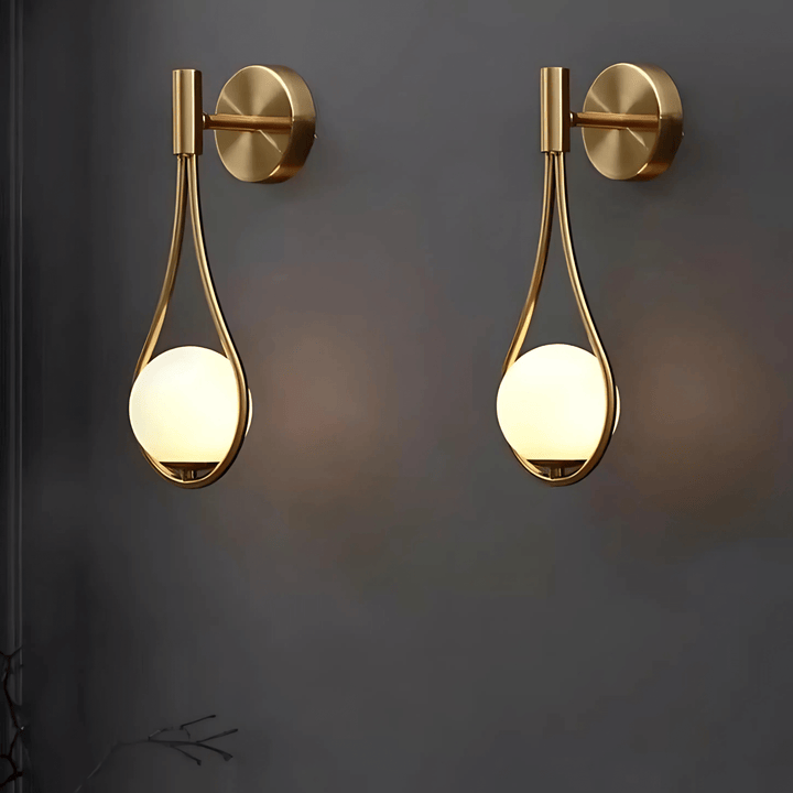 Two modern gold teardrop wall sconces with glowing white globes mounted on a dark gray wall.