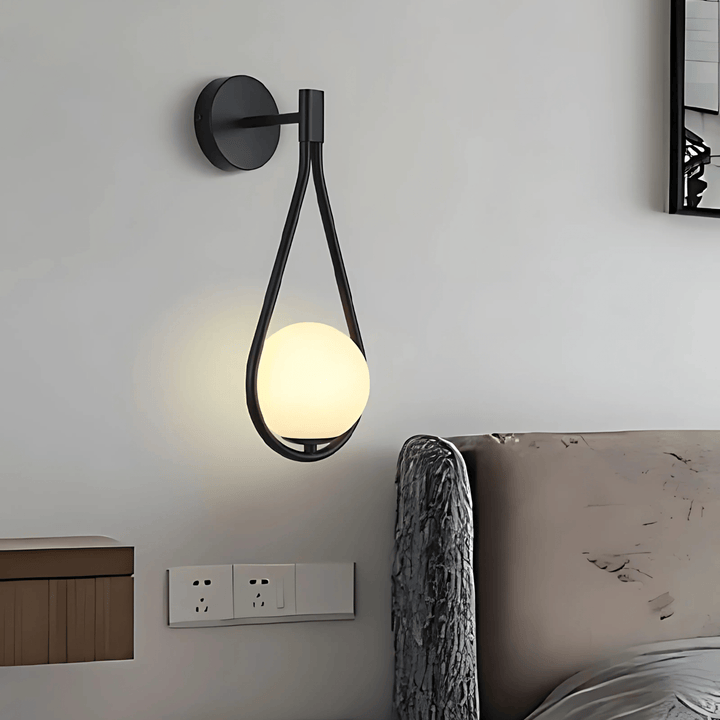 Black teardrop wall sconce with a frosted glass globe light, mounted on a white wall next to a bed.