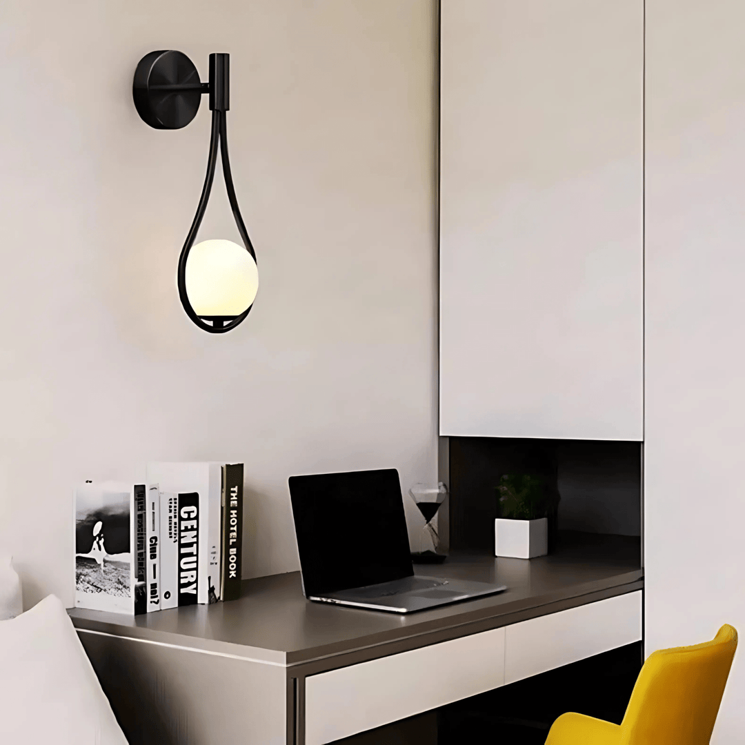 Black teardrop wall sconce with a glowing white orb hanging inside, mounted on a light wall above a desk.