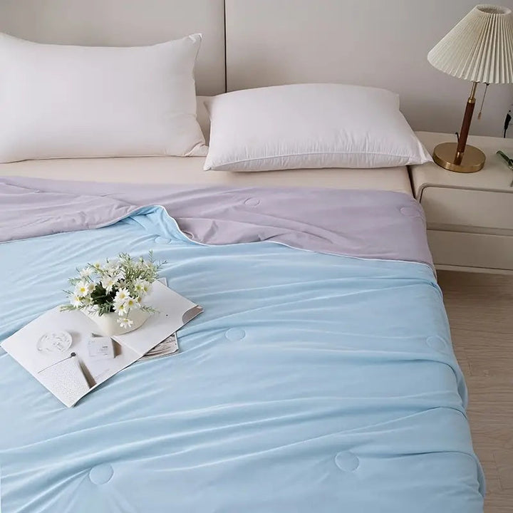 A light blue cooling blanket with a lavender accent is draped over a bed with two white pillows.