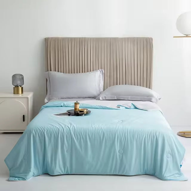 A light blue Forevercool cooling blanket is draped over a bed with grey pillows and a pleated headboard.