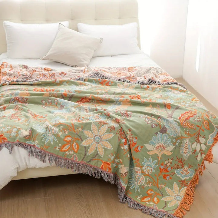 Green cotton blanket with a rustic floral pattern and fringed edges, draped on a bed.