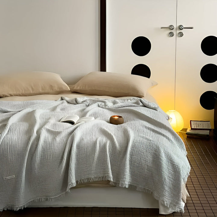 A light grey cotton crinkle tassel blanket is draped over a bed with two beige pillows.