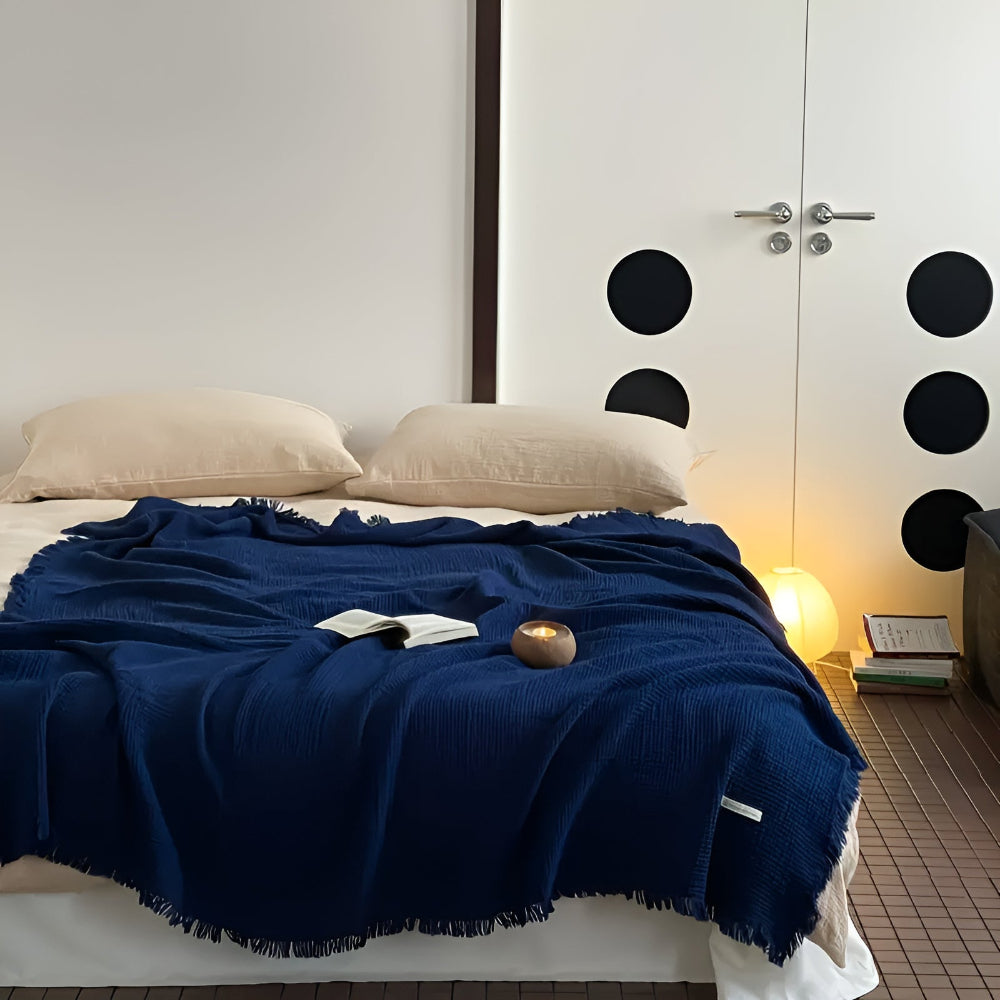 A navy blue cotton crinkle tassel blanket is draped over a bed with two beige pillows.