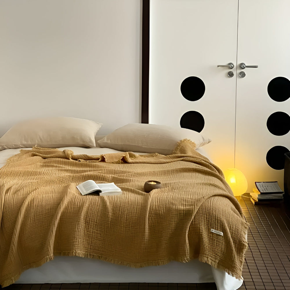 A sand-colored cotton crinkle tassel blanket is draped over a bed with two pillows.