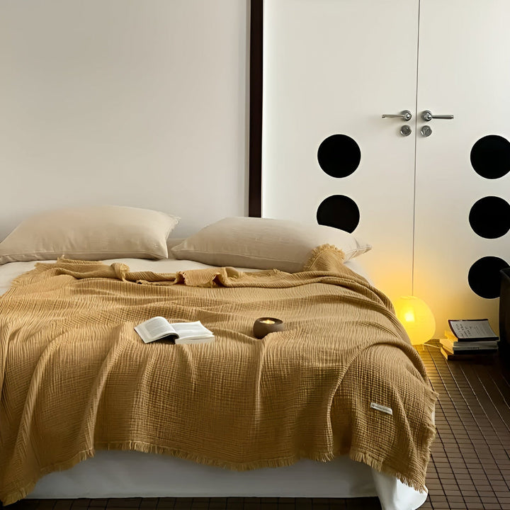 A sand-colored cotton crinkle tassel blanket is draped over a bed with two pillows.