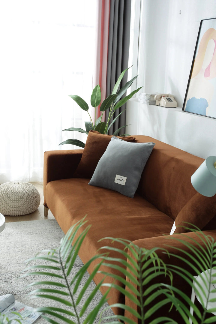 A brown velvet couch cover is shown on a sofa with a grey pillow and a plant.