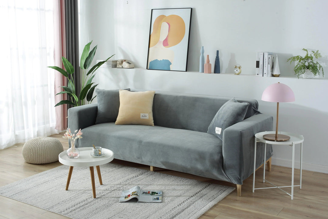 Light grey velvet couch cover on a sofa with decorative pillows and a side table with a lamp.