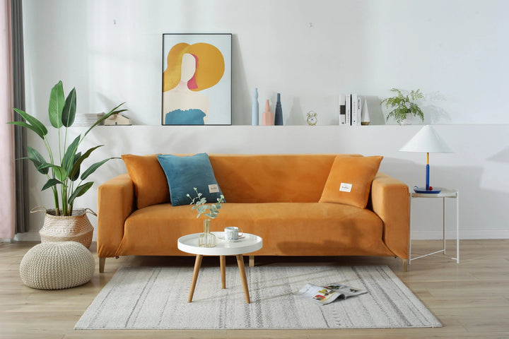 Orange velvet couch cover on a sofa with decorative pillows and a small table.