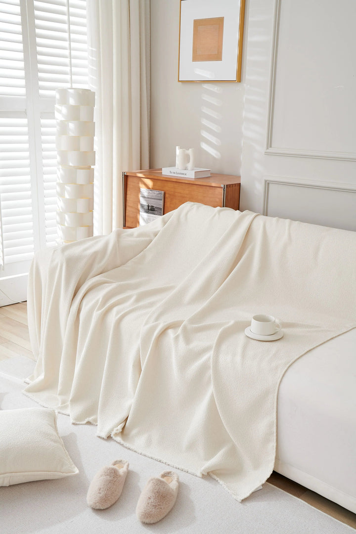 Off-white throw blanket draped over a sofa with a pillow and slippers on the floor.