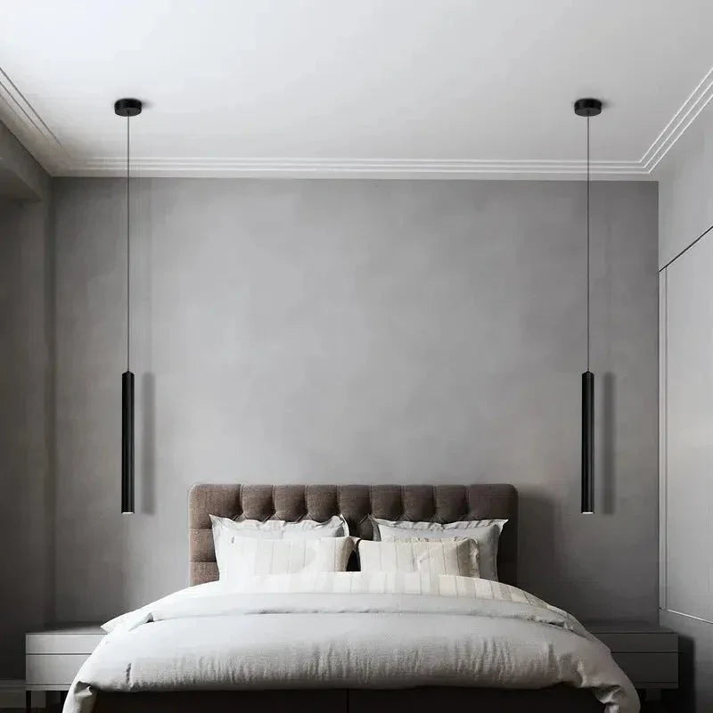 Two black tube pendant lights hanging over a bed with a gray headboard and white bedding.
