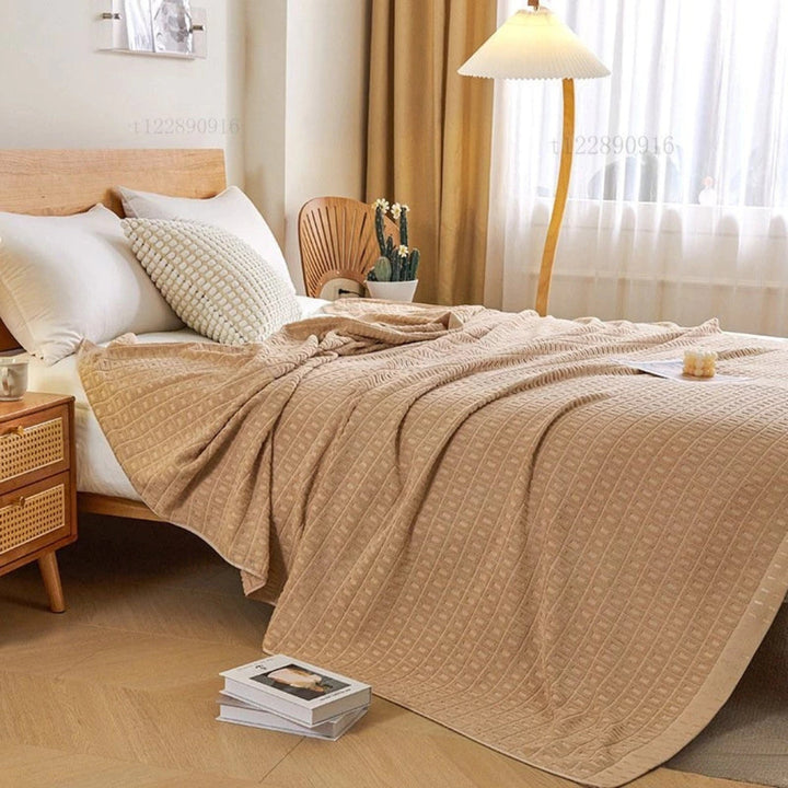 Beige waffle cotton throw blanket draped on a bed with white pillows and a wooden headboard.